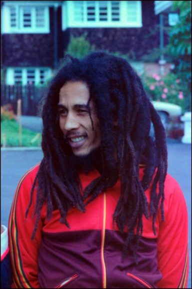 Ein Mann mit Dreadlocks trägt eine rote Jacke und lächelt. Hintergrund mit Gebäuden.